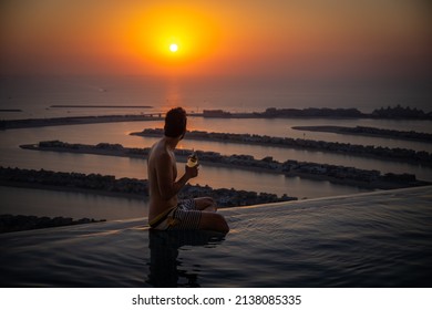 Dubai, UAE - Dec 05 2021: Young Man Enjoying The Beautiful Sunset Panorama With A Cocktail In His Hand On The Edge Of An Infinity Pool, With The Leaves Of Palm Jumeirah In The Background