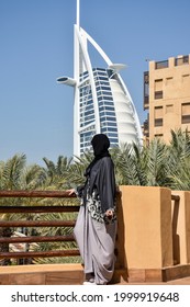 Dubai, UAE - February 18, 2018: Low Angle View Of Young Arab Woman Wearing A Modern Abaya And Facing Burj Al Arab. Fashion Concept.