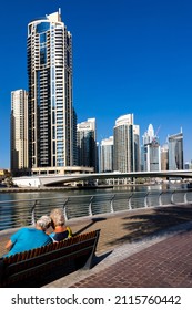 Dubai, UAEmirates - Jan 31 2022: An Elderly Tourist Couple Takes A Rest On One Of The Many Beautiful Benches Along The Southern Promenade Of The Dubai Marina. High Rise Buildings On The Other Side.
