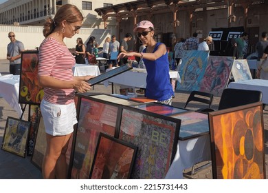 Dubai, United Arab Emirates - 24 October 2015: Woman Holds Framed Artwork At Hip Art Fair In Al Fahidi. Female Artist Behind Table Points And Talks About Her Work. Sunny Outdoor Weekend Summer Event.