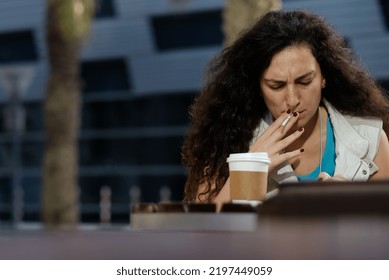 Dubai, United Arab Emirates - February 15, 2017: Woman With Long Curly Hair And Maroon Nails Takes A Cigarette And Coffee Work Break, Midday, Outside A Cafe. Female Looks Down With A Frown.