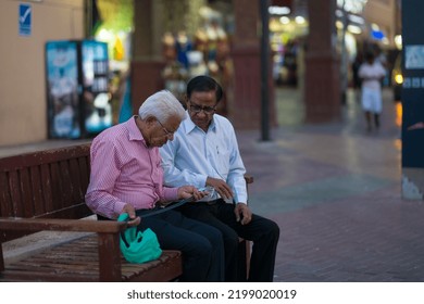 Dubai, United Arab Emirates - February 12, 2018: Two Old Men Wearing Eyeglasses, Button-up Shirts, And Slacks, Sit On A Bench Inspecting A Belt They Just Bought From The Textile Souk (Arabic Market).