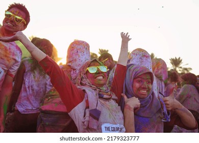 Dubai, United Arab Emirates - November 29, 2013: Young Arab Woman Covered In Colored Powder Raises Both Arms Up In Joy At The Color Walk. She Has A Hijab, Shades, A Huge Smile, And A Female Friend.