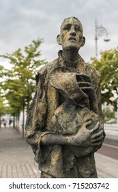 Dublin, Ireland - August 7, 2017: Great Irish Famine Bronze Statue Set On Custom House Quay Along Liffey River In Docklands. One Slender Male Figure. Green Trees And Gray Sky.