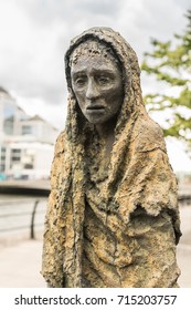 Dublin, Ireland - August 7, 2017: Great Irish Famine Bronze Statue Set On Custom House Quay Along Liffey River In Docklands. One Slender Female Figure. Green Trees And Gray Sky.