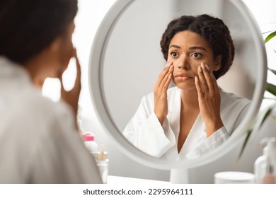 Dull Skin Concept. Upset Black Woman Looking At Mirror And Touching Face, Disappointed Young African American Female Sitting At Dressing Table, Checking Dark Circles Under Eyes And Wrinkles