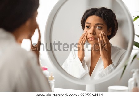 Dull Skin Concept. Upset Black Woman Looking At Mirror And Touching Face, Disappointed Young African American Female Sitting At Dressing Table, Checking Dark Circles Under Eyes And Wrinkles