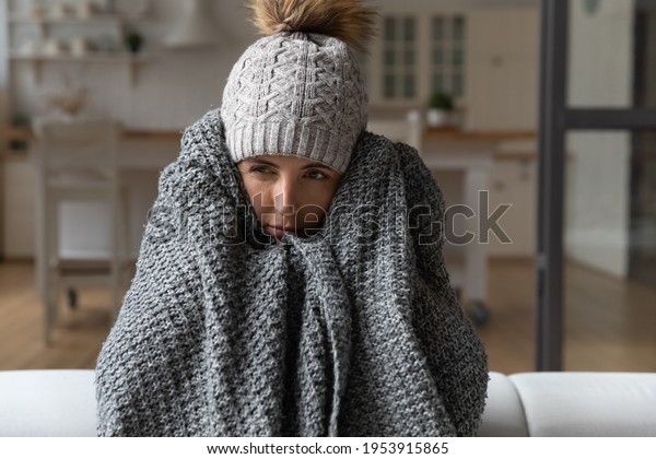 Dull young hispanic woman save herself from freezing\
wear winter clothes muffle up in blanket think of buying radiator\
heater. Shivering young lady sit on sofa in plaid ponder on much\
too cold at home