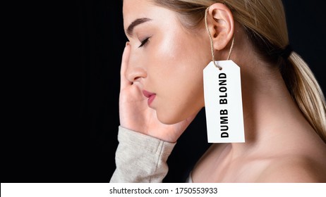 Dumb Blonde. Tired And Sad Woman Touches Temple With Her Hand With Label On Ear Isolated On Black Background, Studio Shot, Cropped