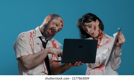 Dumb Creepy Looking Zombies With Modern Laptop Device And Clipboard Acting Childish On Blue Background. Sinister Looking Brain-eating Monsters With Deep Scars And Wounds Working On Portable Computer.