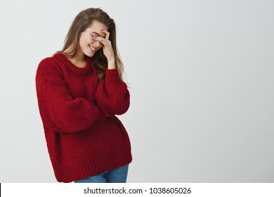 So Dumb But So Sweet. Studio Shot Of Attractive Stylish Woman In Red Loose Sweater Making Facepalm, Holding Hand On Eyes And Smiling, Coming To Terms With Stupid Sense Of Humour Of Boyfriend