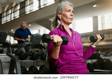 Dumbbell, fitness and senior woman at a gym for training, wellness and cardio with earphones, music or mindset. Weightlifting, bodybuilding and elderly female person at sports center for arm workout - Powered by Shutterstock