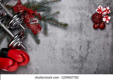 Dumbbells, Boxing Gloves,  Fir Tree Branches And Christmas Decorations On A Gray Background.Top View With Copy Space. New Year And Christmas. Fitness, Sport And Healthy Lifestyle Concept.