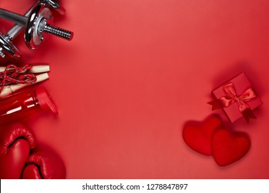 Dumbbells, boxing gloves, rope, bottle for water, gift box and two red hearts on a red background.Top view with copy space. Valentine's Day card. Fitness, sport and healthy lifestyle concept.