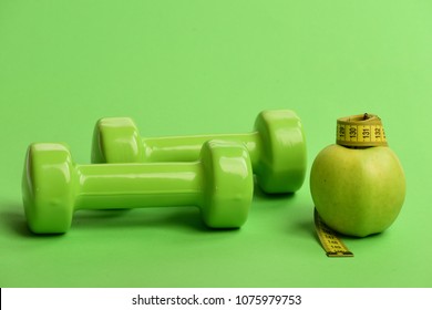 Dumbbells In Bright Green Color, Measure Tape And Fruit On Green Background. Sports And Healthy Regime Symbols. Tape Measure In Yellow Color By Barbells And Juicy Apple. Diet And Sport Regime Concept