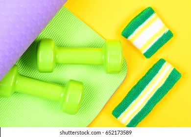 Dumbbells on green and purple mat for yoga and sweatband with wristband isolated on yellow background, top view