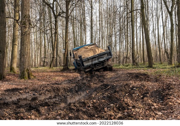 Dump truck car stuck in mud on the road\
among trees. Blurry mud bad road in the park. Тruck dump car with\
sand stopped due to wet dirty on its way to\
park.