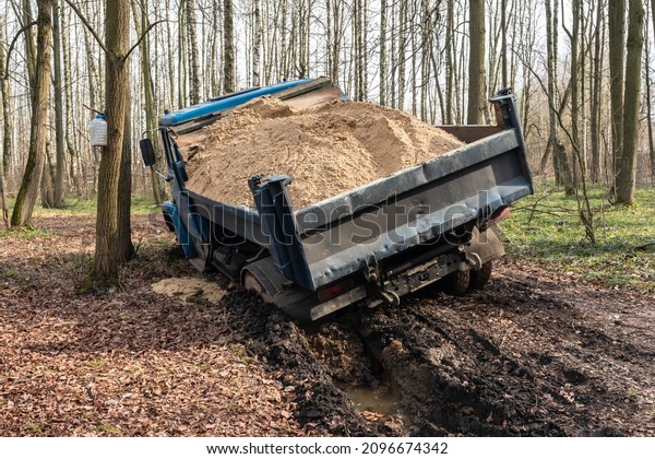 Dump truck car stuck in mud on the road\
among trees. Blurry mud bad road in the park. Тruck dump car with\
sand stopped due to wet dirty on its way to\
park.
