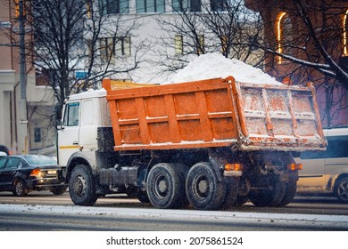 Dump Truck Loaded With Snow Moving On City Road In Evening During Snow Storm. Utility Service Removing Snow From City Streets, Cleaning Streets At Winter Season