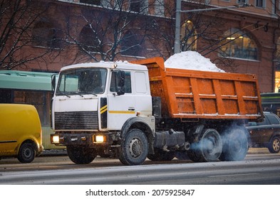 Dump Truck Loaded With Snow Moving On City Road In Evening During Snow Storm. Utility Service Removing Snow From City Streets, Cleaning Streets At Winter Season.