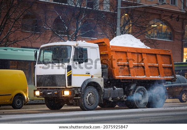 Dump truck loaded with snow\
moving on city road in evening during snow storm. Utility service\
removing snow from city streets, cleaning streets at winter\
season.