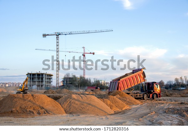 Dump truck unloading earth sand for road\
construction or for foundation work. Work of tower crane at\
construction site on blue sky background. Tall house renovation\
project, government\
programs