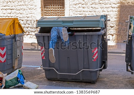 Dumpster diving: man in denim jeans climbs into a large green rubbish (trash) bin in the street; just his feet are sticking out. He retrieves items of perceived value to use or sell  for profit.