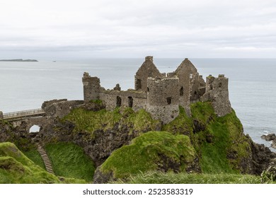 Dunluce Castle sits on a unique rocky cliff overlooking the ocean, with steep green slopes and rugged terrain. This historic landmark is framed by the sea and cloudy sky - Powered by Shutterstock