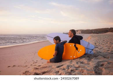 A duo, an adult male and a small boy, sharing a joyful surfing experience sit in wetsuits at a beach under the setting sun - Powered by Shutterstock