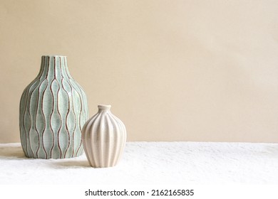 Dúo de jarrones sobre un fondo beige. Un jarrón azul cielo de tamaño mediano con ranuras marrones y un pequeño jarrón beige. 