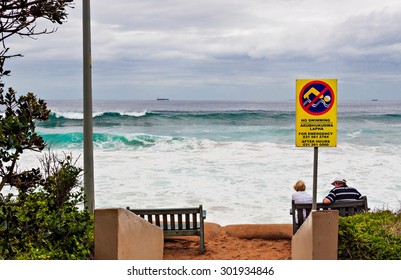 DURBAN, SOUTH AFRICA - January 20, 2015: Man And Woman Sitting On A Bench In Front Of A No Swimming Sign Along The Promenade At The Beach In Umhlanga Rocks