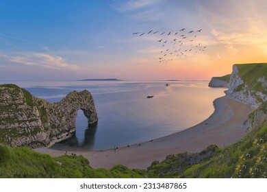 Durdle door Beach with a flock of birds in the purple and magenta sunset sky and a silhouette of a paddle boat in the sea and natural stone arch in the foreground - Powered by Shutterstock