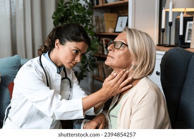 During a home visit to an elderly patient, a young endocrinologist doctor checks her thyroid gland by feeling her neck with her hands. Senior women endocrinology health care - Powered by Shutterstock