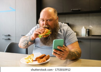During lunch break. Bearded tattooed involved man eating appetite burger looking at smartphone in kitchen at table