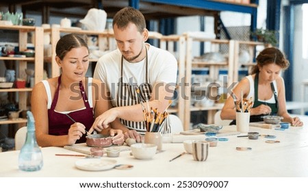 During master class, student paints clay vessel under guidance of male teacher. Mentor is next to student, makes comments and corrects inaccuracies