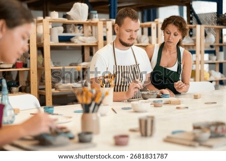 During master class, student paints clay vessel under guidance of female teacher. Mentor is next to student, makes comments and corrects inaccuracies
