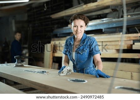 During processing of board preparation materials for execution manufacture of individual orders in carpentry workshop, woman master grinds surface of board, eliminates irregularities, removes sawdust