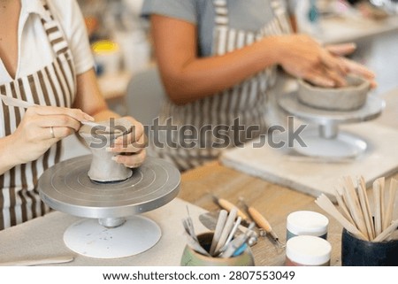 During working day, unrecognizable two pottery workshop female visitors sculpt from raw clay. They smooth surface of product with their fingers, form final product, and carry out order at desk.