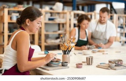 During workshop at master class, female employee paints clay vessel, applying pigment to plate with brush. In background, mentor teaches student, helps to color bowl. - Powered by Shutterstock