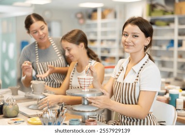 During workshop at master class, female pupil paints clay vessel, applying pigment to plate with brush. In background, mentor teaches student, helps to color bowl. - Powered by Shutterstock