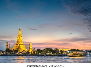 At dusk,the silhouette of a Thai Long Tail boat drifting past the golden pagodas of the temple complex,as they point towards clear sunset skies,seen from opposite side of Bangkoks main river. - Shutterstock ID 2260281413
