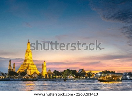 At dusk,the silhouette of a Thai Long Tail boat drifting past the golden pagodas of the temple complex,as they point towards clear sunset skies,seen from opposite side of Bangkoks main river.