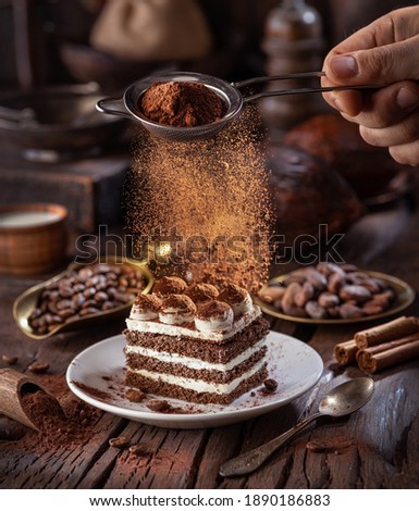 Dusting tiramisu - like cake with cocoa powder. Still life with slice of cake and coffee and cacao beans on wooden table.