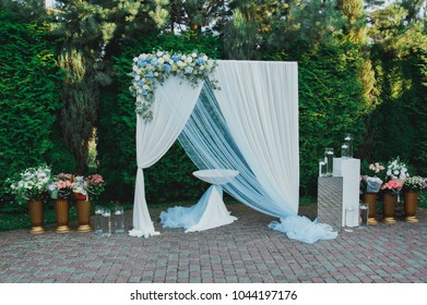 Dusty Blue Wedding Arch Outdoors In The Green Park. Candles With Vases Of Flowers On Background. White And Blue Decorating With Hydrangea Flowers.