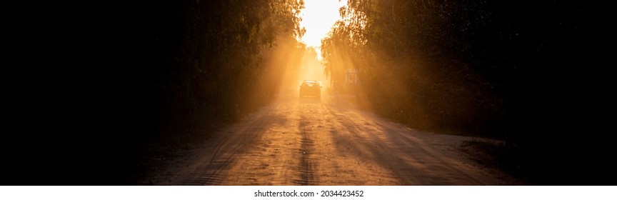 dusty dirt road on a beautiful sunset behind the forest, in the front and background silhouettes of cars