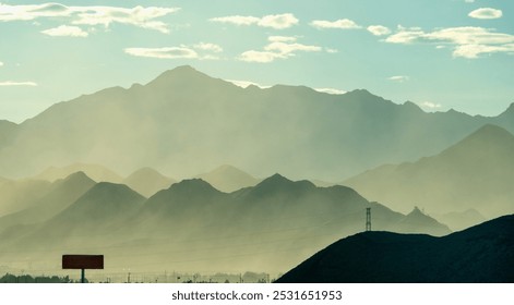 Dusty mountains layered under a bright sky with scattered clouds, a silhouetted tower and sign add depth to the serene landscape. - Powered by Shutterstock