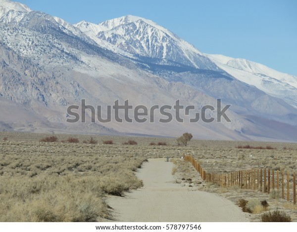 Dusty unpaved road with fence surrounded by desert\
landscape. A mountain peak covered with snow and ice in the\
background under blue sky