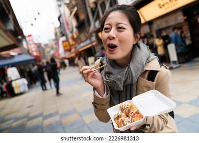 dutch angle shot of cheerful Asian Japanese girl making funny face expression while eating hot delicious takoyaki octopus balls at a food plaza in Shinsekai Osaka Japan