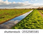 Dutch polder landscape with a straight ditch. It is a windless day in the autumn season and the clouds are reflected in the mirror-smooth water surface.