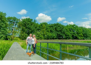 Dutch rural landscape with a walking senior couple with their brown dog on a metal bridge against lush green trees, a sunny spring day to enjoy nature, casual clothes, blue jeans and a white blouse - Powered by Shutterstock